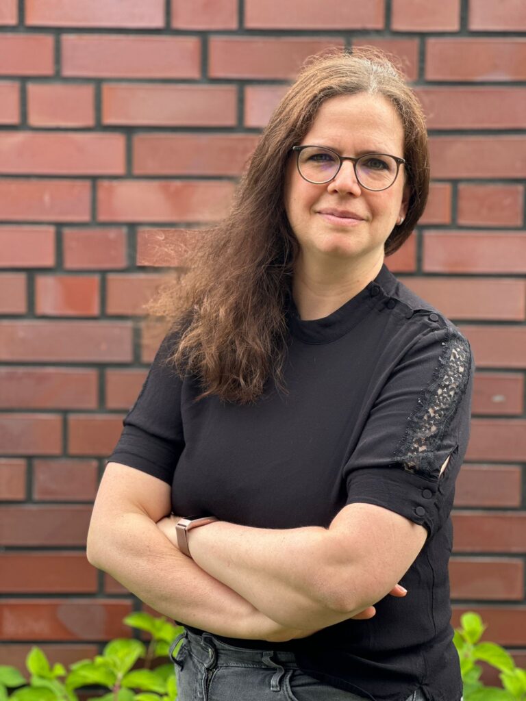 Marketingberatung am Niederrhein: Melina Kilzer mit offenen Haaren und einer schwarzen, kurzärmeligen Bluse vor einer Backsteinwand
